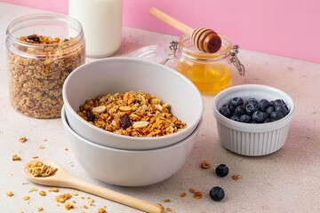 Delicious granola with raisin, sunflower seeds, peanut and hazelnut in bowl served with bottle of milk, honey and blueberries on pink background. Healthy vegan breakfast food. Selective focus.