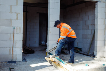 Mature worker in orange protect coveralls sawing wood and metal tubes near wall. Laying foundation....