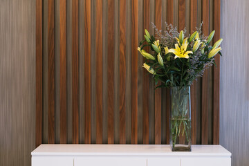 Yellow lily flower in square glass vase on the white cabinet with wooden stripe background