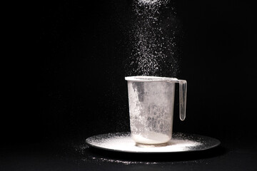 White wheat flour is poured into a measuring cup on a black background. Cooking and baking, a place for text.