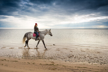 wearing jacket, jeans and warm hat girl horses a gray palfrey along shore