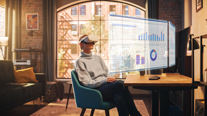 Young Female Employee with Short Hair Works From Home in Virtual Reality Goggles. She is Wearing a Modern Headset and Using Controllers to Scroll Through Graphs. Stylish Apartment with Big Window.