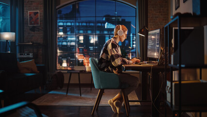 Young Woman Writing Code on Desktop Computer in Stylish Loft Apartment in the Evening. Creative Female Wearing Headphones, Working from Home on Software Development. Urban City View from Big Window. - Powered by Adobe