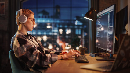 Young Woman Writing Code on Desktop Computer in Stylish Loft Apartment in the Evening. Creative Female Wearing Headphones, Working from Home on Software Development. Urban City View from Big Window.