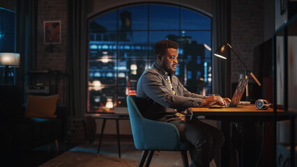 Young Handsome African American Man Working from Home on Laptop Computer in Stylish Loft Apartment at Night. Creative Male Checking Social Media, Browsing Internet. Urban City View from Big Window.