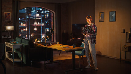 Young Beuatiful Women Working from Home, Standing And Holding Laptop Computer in Stylish Loft Apartment in the Evening. Creative Female Wearing Cozy Casual Outfit. Urban City View from Big Window.