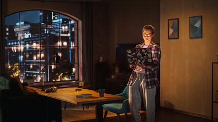Fototapeta na wymiar Young Beuatiful Women Working from Home, Standing And Holding Laptop Computer in Stylish Loft Apartment in the Evening. Creative Female Wearing Cozy Casual Outfit. Urban City View from Big Window.