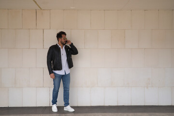 Handsome young man with beard, sunglasses, leather jacket, white shirt and jeans, next to a white wall, talking on his cell phone. Concept beauty, fashion, trend, smartphone, app, social networks.