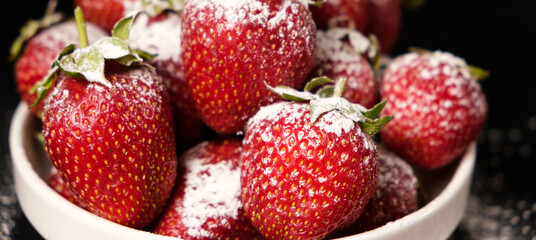 Strawberries in a plate with powdered sugar on a black background, side view close-up.