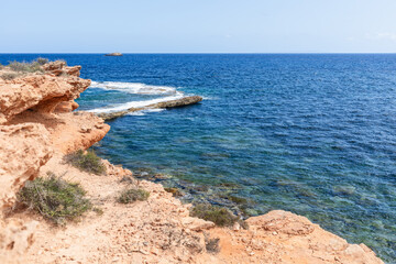 Orange-pink rocky coast with rare plants, water changes color depending on depth, clear sky, summer day, Ibiza, Balearic Islands, Spain