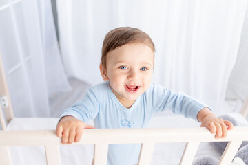 a happy baby boy in a crib in the children's room is holding on to the side and smiling