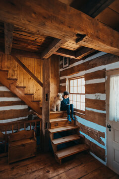 Boy and dog sitting on stairs of log cabin home looking out window.