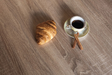 A cup of hot fragrant black coffee in a saucer with croissant and cinnamon sticks, on a light brown wooden table.