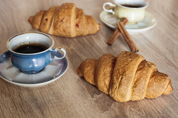 A couple of cups of hot fragrant black coffee in a saucer with croissants and cinnamon sticks on a light brown wooden table.