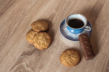 A cup of hot fragrant black coffee in a saucer with cookies and a chocolate bar, on a wooden light brown textured table.