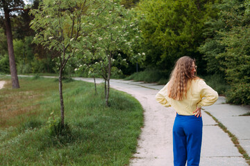 A smilling girl with blond curly hair dressed in blue jeans and a yellow shirt, wearing sunglasses, stands on a path in a picturesque park in summer.