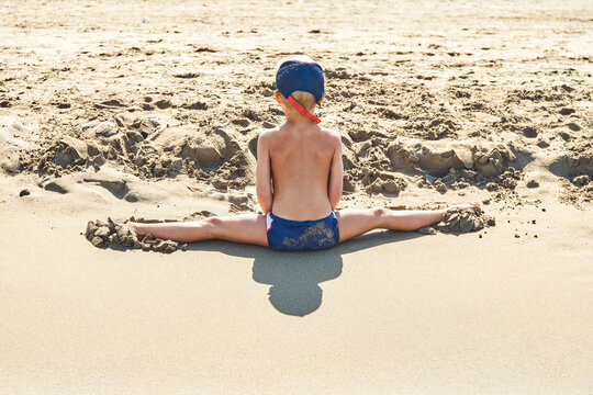 Preschooler makes leg-split on soft sand of seashore. Little boy in sun protective hat sits on beach enjoying summer vacation backside view