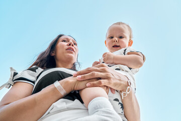 Mother holds cute smiling toddler in arms against blue sky. Young brunette woman and little boy...
