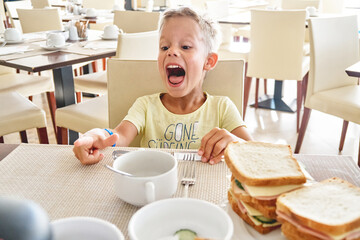 Blond preschooler boy laughs while having breakfast at hotel. Cute boy has fun sitting at table and eating big sandwiches on summer vacation