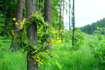 wreath of wildflowers hanging on tree in forest, natural abstract green background. wreath - floral traditional decor, symbol of Summer Solstice Day, Midsummer. pagan witch traditions, wiccan ritual. - obrazy, fototapety, plakaty