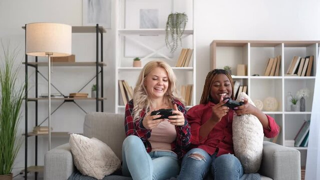 Fun entertainment. Friends home leisure. Weekend joy. Excited girl cheering up bored bff offering to play video game together on couch in modern living room interior.