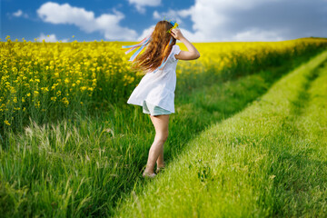 Teenage girl in white dress and Ukrainian wreath runs through yellow fields and green meadows, against cloudy sky