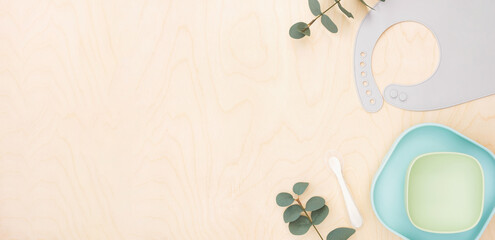 Baby accessories and tableware for food on wooden background. First feeding for kids concept. Flat lay, Top View, Space for text. Silicone dishware, bib, cup on neutral backdrop. Serving baby food