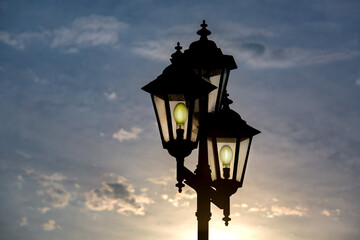 Fototapeta na wymiar silhouette retro style triple lantern head with transparent glass panels with glow electric light bulbs, iron street lighting in the evening at sunset against a blue sky with sun light.