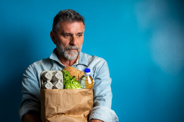 Surprised mature man looking at store receipt after shopping, holding a paper bag with healthy food. Real people expression. Inflation concept. man with a paper bag of groceries looks surprised.