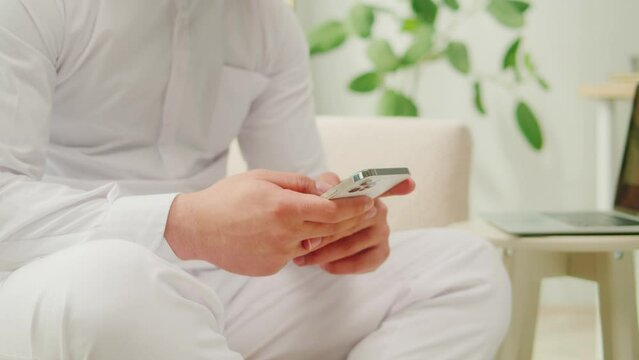 Middle eastern man using smartphone. Male person texting at phone in living room. Wearing traditional Islamic clothes, relaxing at home. Communicating with family and friends online.