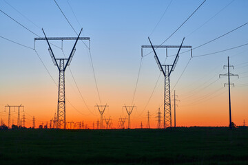 Dark silhouettes of power lines on fiery sunrise. High voltage electricity towers in field and fiery sunset. Concept of crisis of energy consumption, generation, and supply.