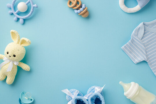 Baby accessories concept. Top view photo of infant clothes blue shirt knitted booties bunny toy teether rattle bottle and pacifier on isolated pastel blue background with empty space in the middle