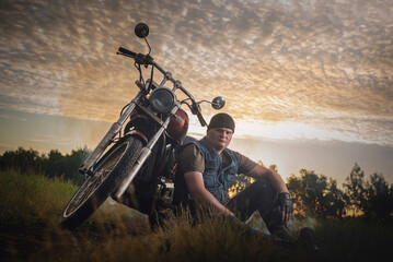 Motorbiker sits near the motorbike on the dusty empty countryside road at the sunset sun...