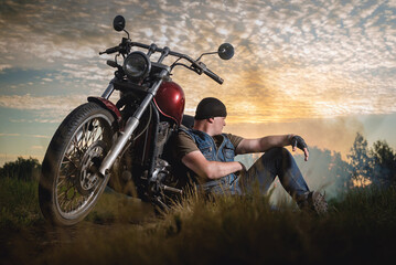 Motorbiker sits near the motorbike on the dusty empty countryside road at the sunset sun...