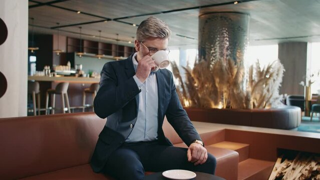 Middle-aged Caucasian businessman sitting at table in cafe, looking at watch. Handsome gray-haired man drinking coffee while waiting for meeting with business partner. Indoor. Beautiful interior in