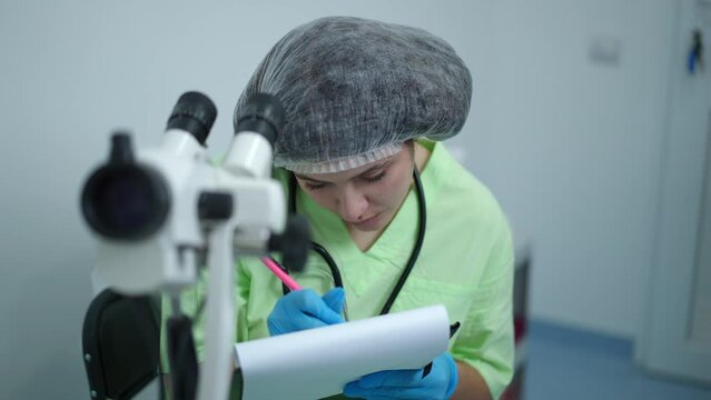 Concentrated expert obstetrician using gynecological colposcope writing with pen thinking. Portrait of expert Caucasian focused gynecologist doing examination in hospital indoors