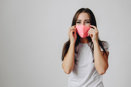 brunette in pink mask on white background smiles joyfully. girl adjusts protective mask on her face
