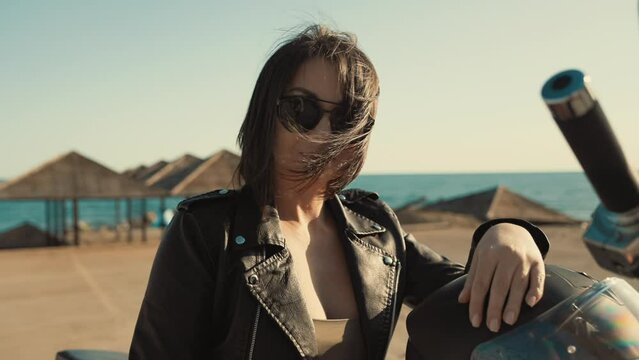 Portrait of a self-confident adult woman in glasses and a leather jacket posing on a motorcycle. Pan shot. Slow motion. The concept of girl power and freedom.
