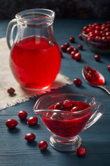 There is a gravy bowl with cranberry sauce on a blue wooden table. Next to it are cranberries, in the background a small jug of cranberry juice.