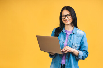 Portrait of happy young beautiful surprised woman with glasses standing with laptop isolated on yellow background. Space for text. - obrazy, fototapety, plakaty