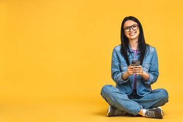 Business concept. Portrait of happy young woman in casual sitting on floor in lotus pose and holding mobile phone isolated over yellow background.