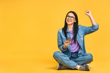 Happy winner! Business concept. Portrait of happy young woman in casual sitting on floor in lotus pose and holding mobile phone isolated over yellow background. Celebrating victory.