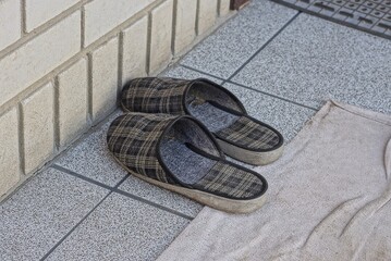 two black cloth slippers stand on a stone floor and a gray rug against a white brick wall outside