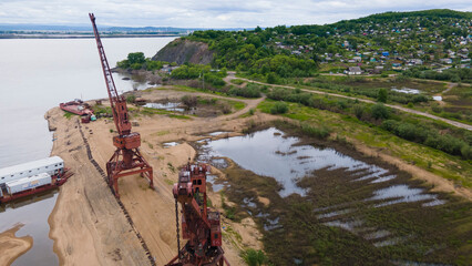 Aerial view of an old abandoned burgundy cargo ship cranes stands at the bank of a river in a hollow near a village on the hills on a sunny summer day, photo