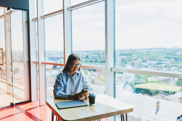 Successful woman is looking at a laptop in a cafe and drinking coffee. A young smiling woman in glasses sits at a table near the window with a phone. Freelance and remote work. Modern female lifestyle