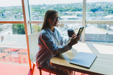 Successful woman is looking at a laptop in a cafe and drinking coffee. A young smiling woman in glasses sits at a table near the window with a phone. Freelance and remote work. Modern female lifestyle