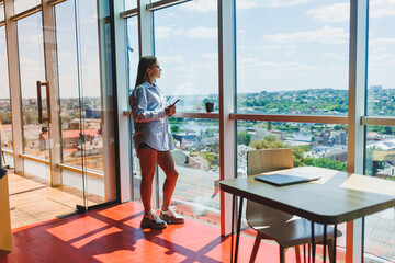 Fototapeta premium Attractive female blogger with a modern smartphone in her hands stands at the window, thoughtful look and daydreaming, holding a mobile phone, ponders an idea for a publication in social networks
