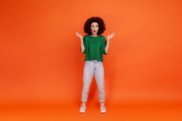 Full length portrait of surprised woman with Afro hairstyle wearing green casual style sweater standing with raised arms, shocking news. Indoor studio shot isolated on orange background.