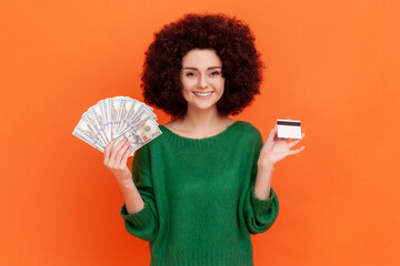 Satisfied woman with Afro hairstyle wearing green casual style sweater showing to camera dollar banknotes and credit card, investments. Indoor studio shot isolated on orange background.