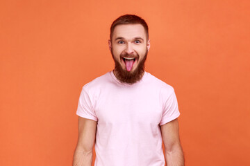 Portrait of bearded man showing out tongue and looking at camera with naughty disobedient grimace, making face, wearing pink T-shirt. Indoor studio shot isolated on orange background.